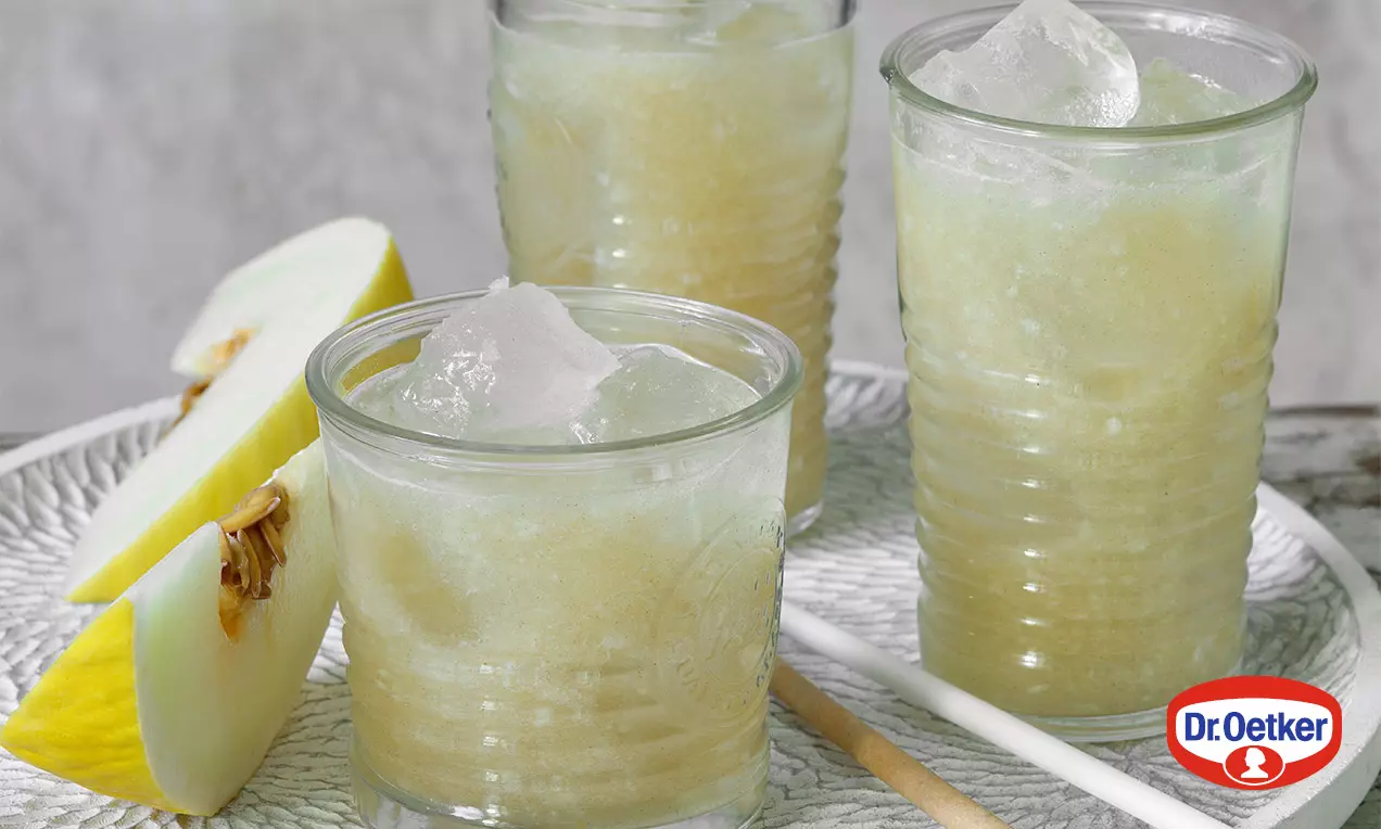 Svetlozelené melónové smoothie s ľadovými kockami v rebrovaných sklenených pohároch, na bielom podnose s kúskami žltého melóna