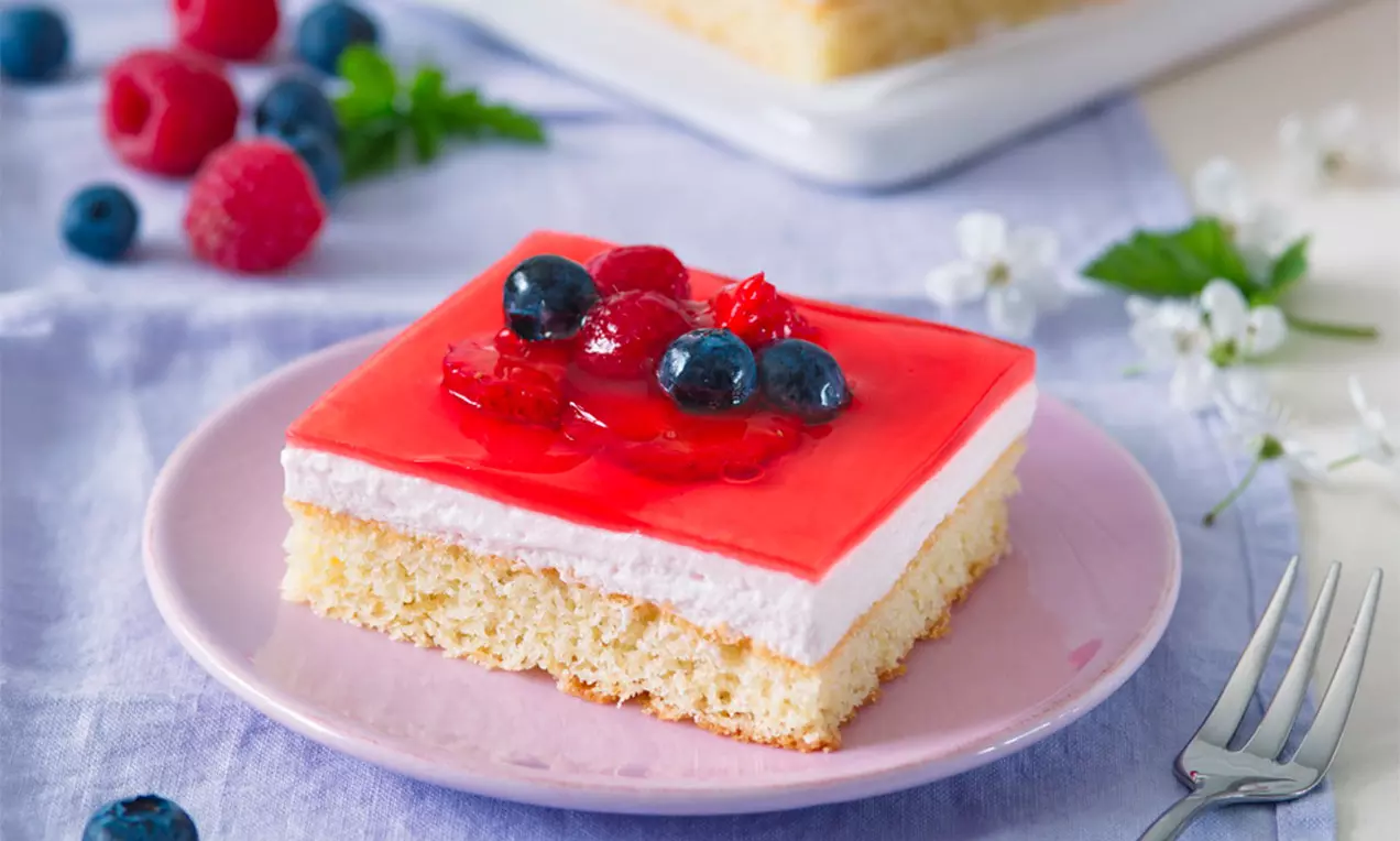 Jemný žltkastý korpus s vrstvou bieleho krému, pokrytý lesklým červeným želé, ozdobený lesným ovocím na pastelovom ružovom tanieri