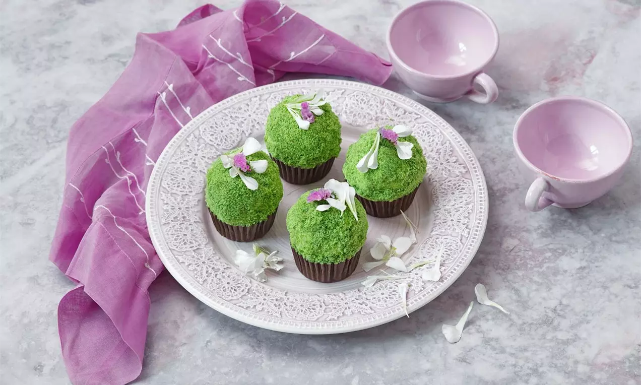 Cupcakes pokryté sýtozelenou posýpkou, ozdobené jemnými kvietkami na zdobenom tanieri, vedľa ružovej šatky a porcelánových šálok