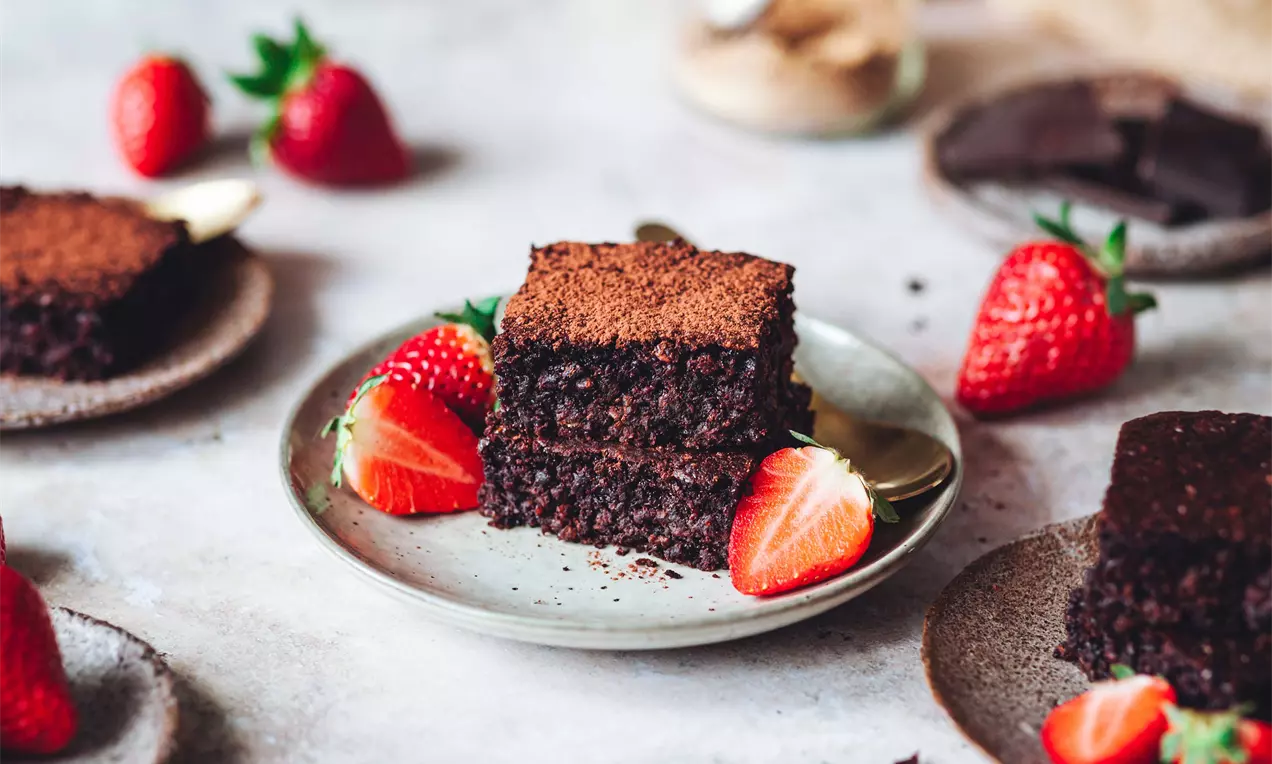 Dva kvádre tmavohnedého ovseno-čokoládového brownies s jemnou kakaovou posýpkou, uložené na tanieri so zrelými jahodami a lyžičkou