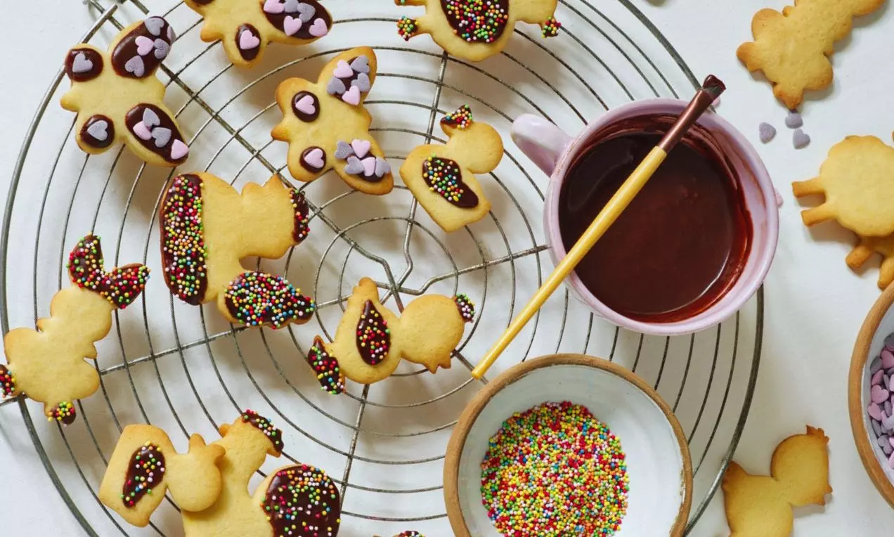 Veľkonočné sušienky zlatisté a chrumkavé, v tvare zvieratiek, zdobené farebnými posýpkami a čokoládou na kovovej mriežke s miskami na zdobenie