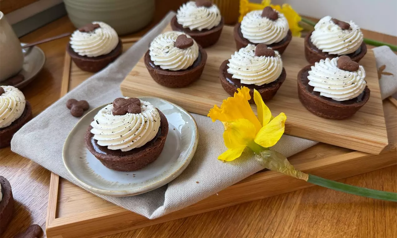 Čokoládové košíčky s krémovo bielou šľahačkou, zdobené malým sušenkovým srdiečkom, na drevenom podnose vedľa žltého kvetu narcisu