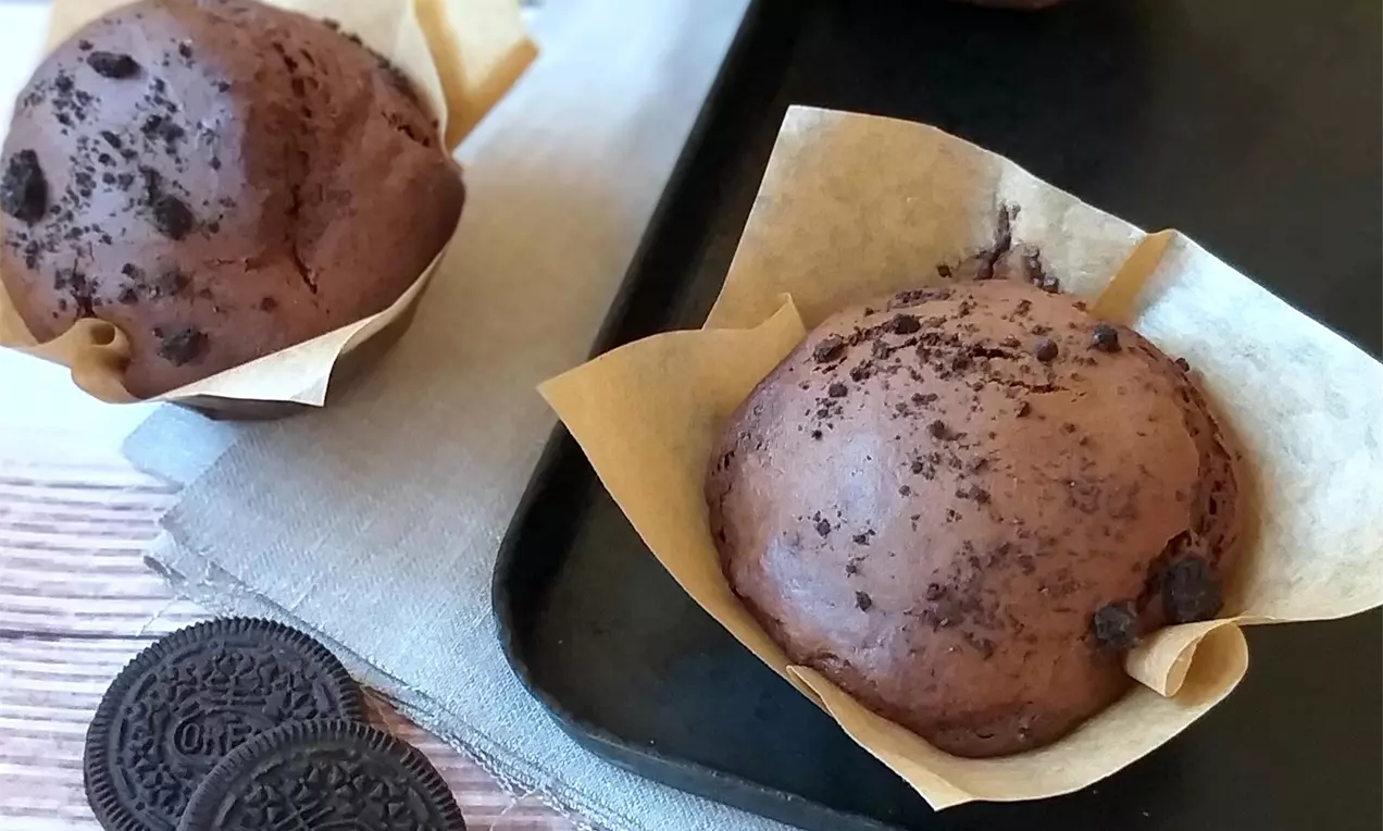 Čokoládové muffiny tmavohnedej farby s kôrkou posypanou kúskami Oreo sušienok, uložené v zlatistých papierových košíčkoch na tmavom plechu