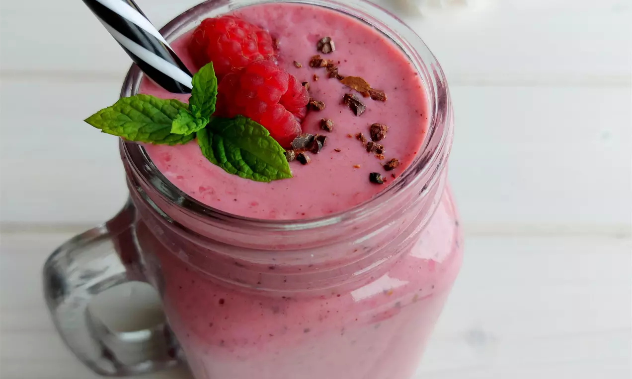 Krémové malinové smoothie v ružovkastom odtieni v priehľadnom pohári, ozdobené čerstvými malinami, lístkami mäty a kakaovými bobmi