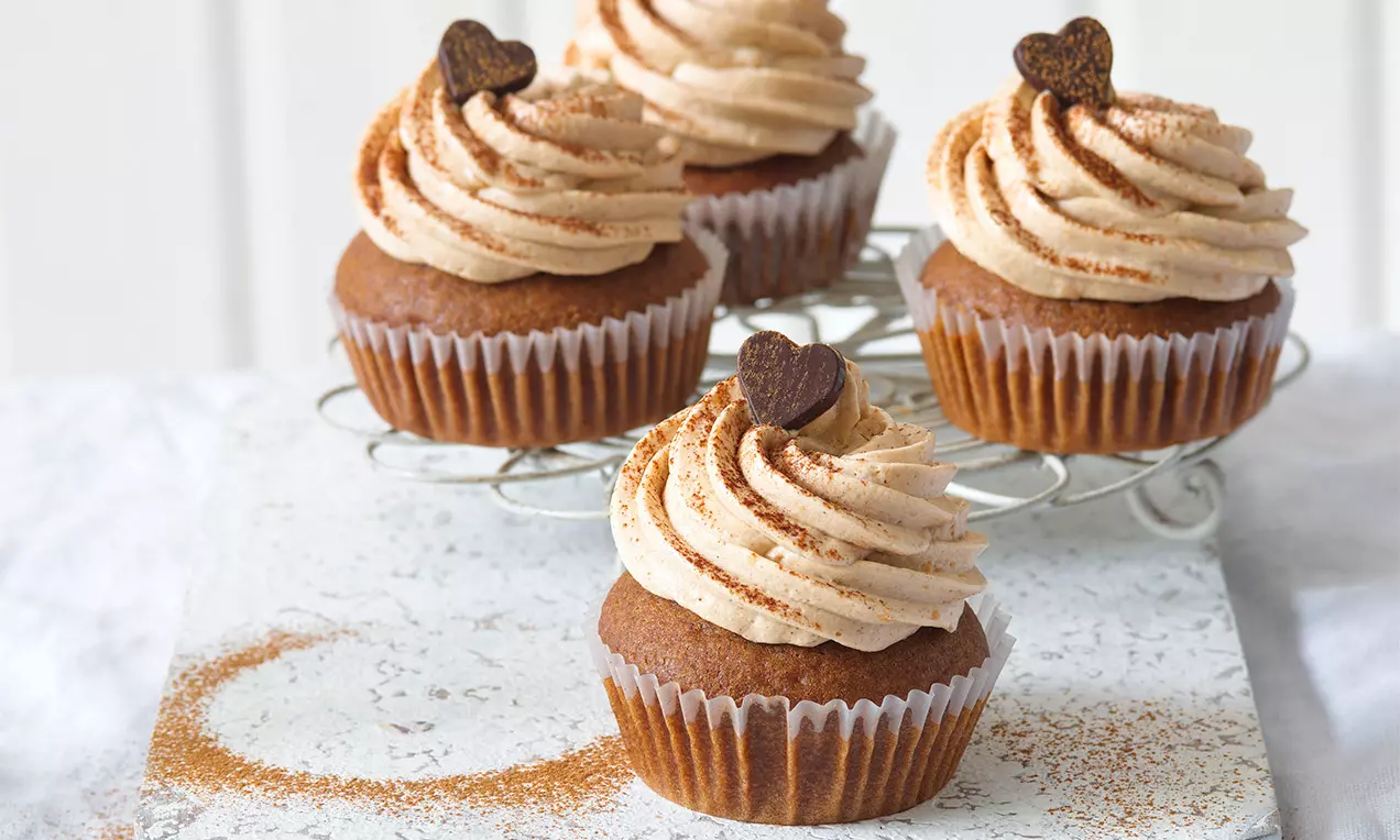 Perníkové cupcakes s nadýchaným krémom svetlohnedej farby, ozdobené čokoládovými srdiečkami a jemným posypom škorice, na mriežke.