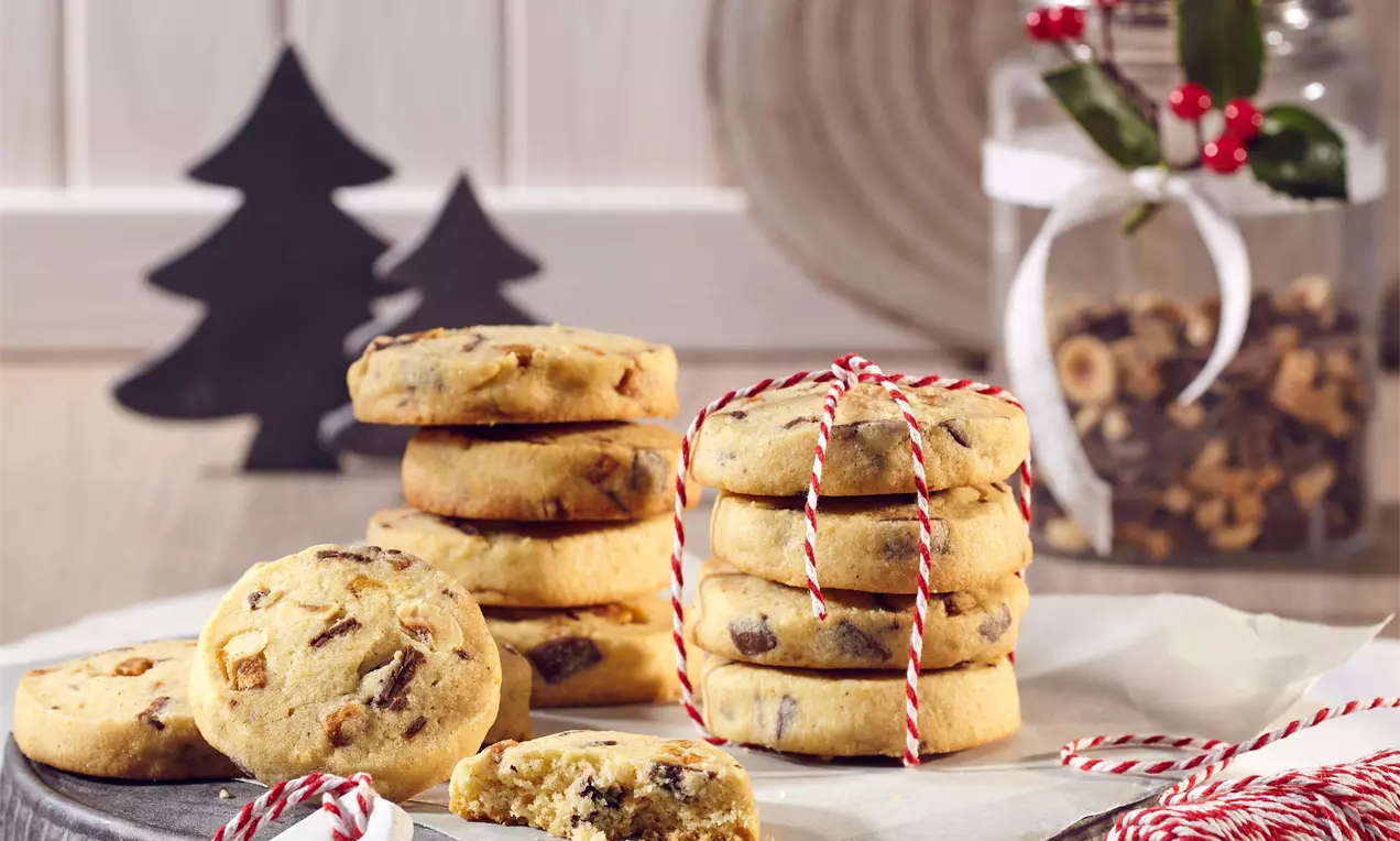 Cookies svetlej zlatohnedej farby s kúskami orieškov a karamelu, naaranžované na papieri s červenobielou šnúrkou, v pozadí sklenený pohár a dekorácie