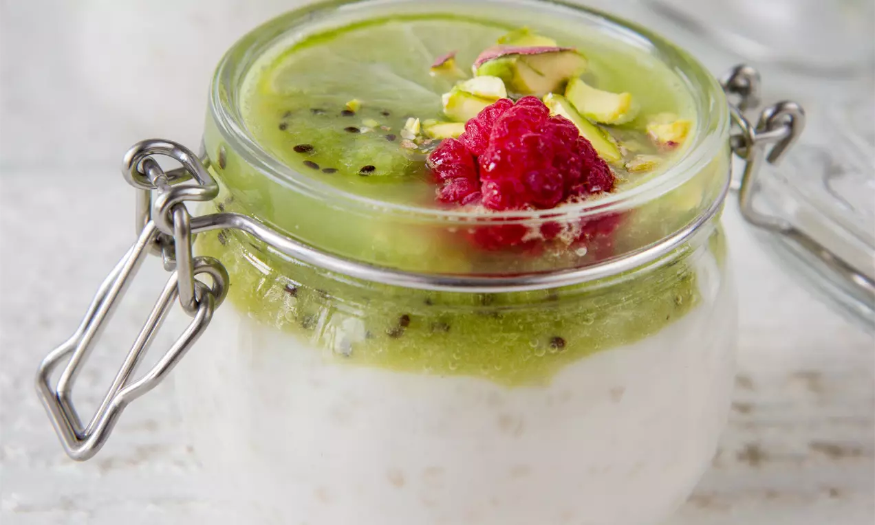 Biely krémový puding v sklenenom pohári s vrstvou zeleného kivi, posypaný kúskami pistácií a červenými malinami