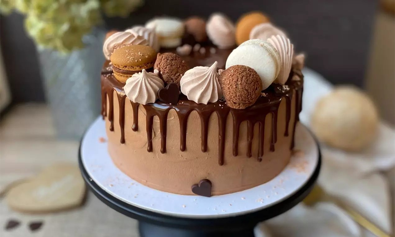 Čokoládová torta s hnedou lesklou polevou stekajúca po bokoch, zdobená makrónkami, krémovými pusinkami a guličkami na čiernom stojane