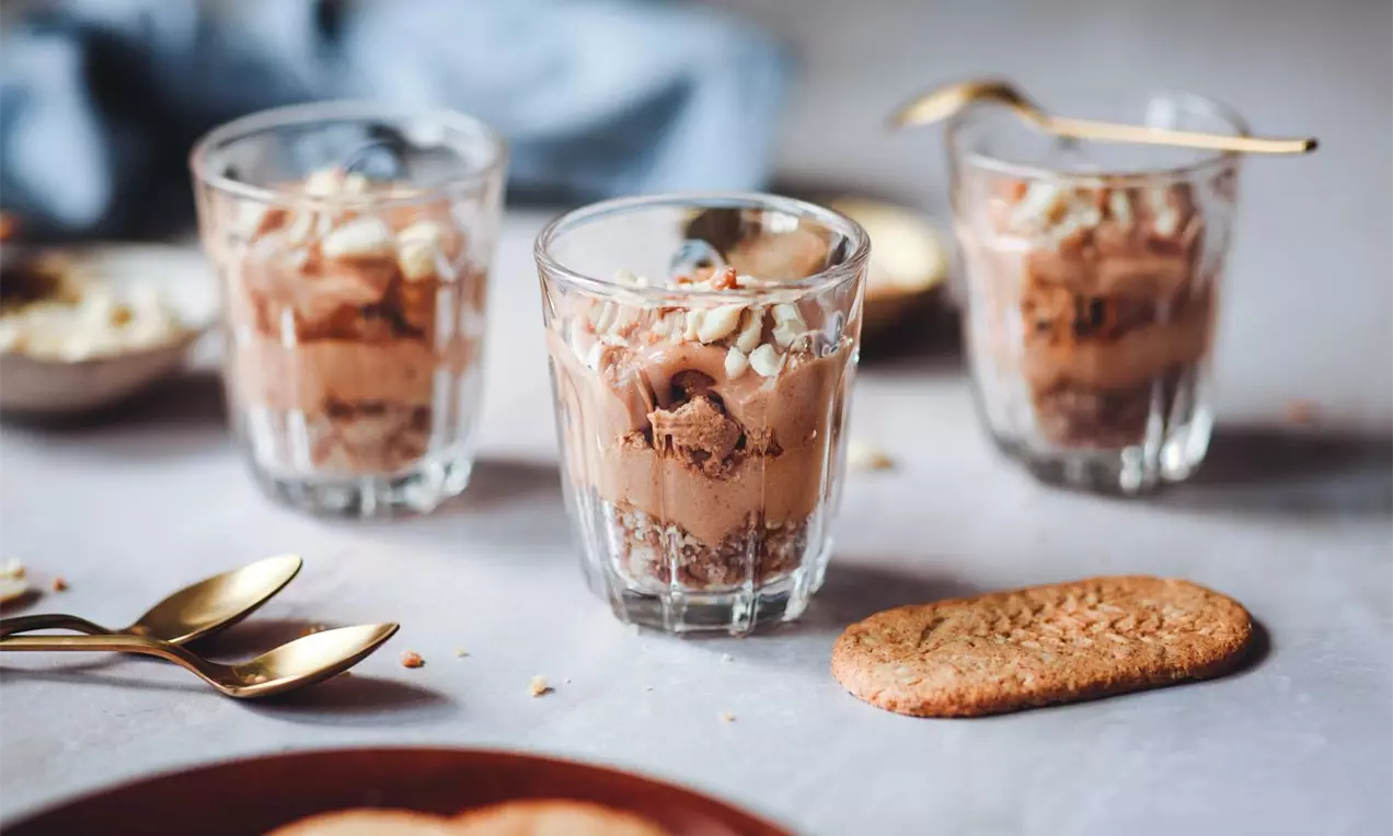 Krémový dezert s čokoládovým mousse v pohároch, zdobený nasekanými arašidmi, so zlatými lyžičkami a sušienkami na podklade.