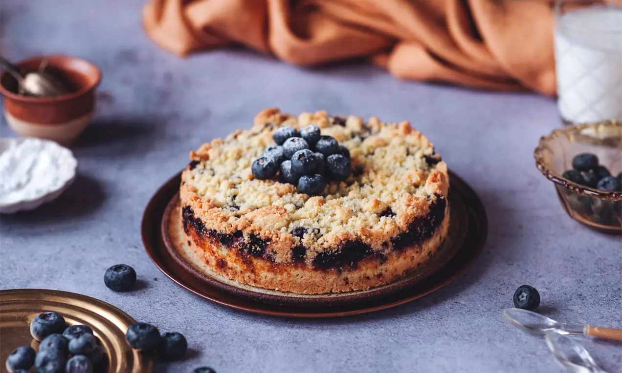 čučoriedkový koláč s posýpkou, ozdobený čučoriedkami, na modrej pracovnej ploche