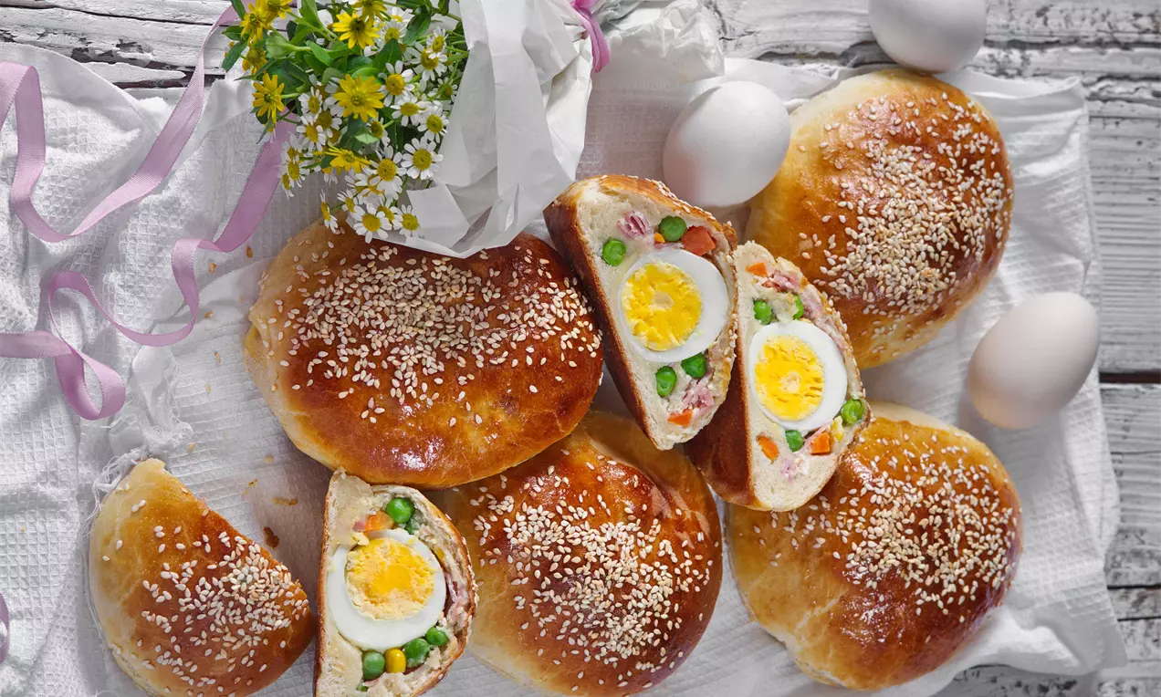 Kynuté houstičky zlatohnedé, posypané sezamom, rozkrojené odhaľujú farebnú náplň s vareným vajíčkom, hráškom a šunkou na bielom obruse