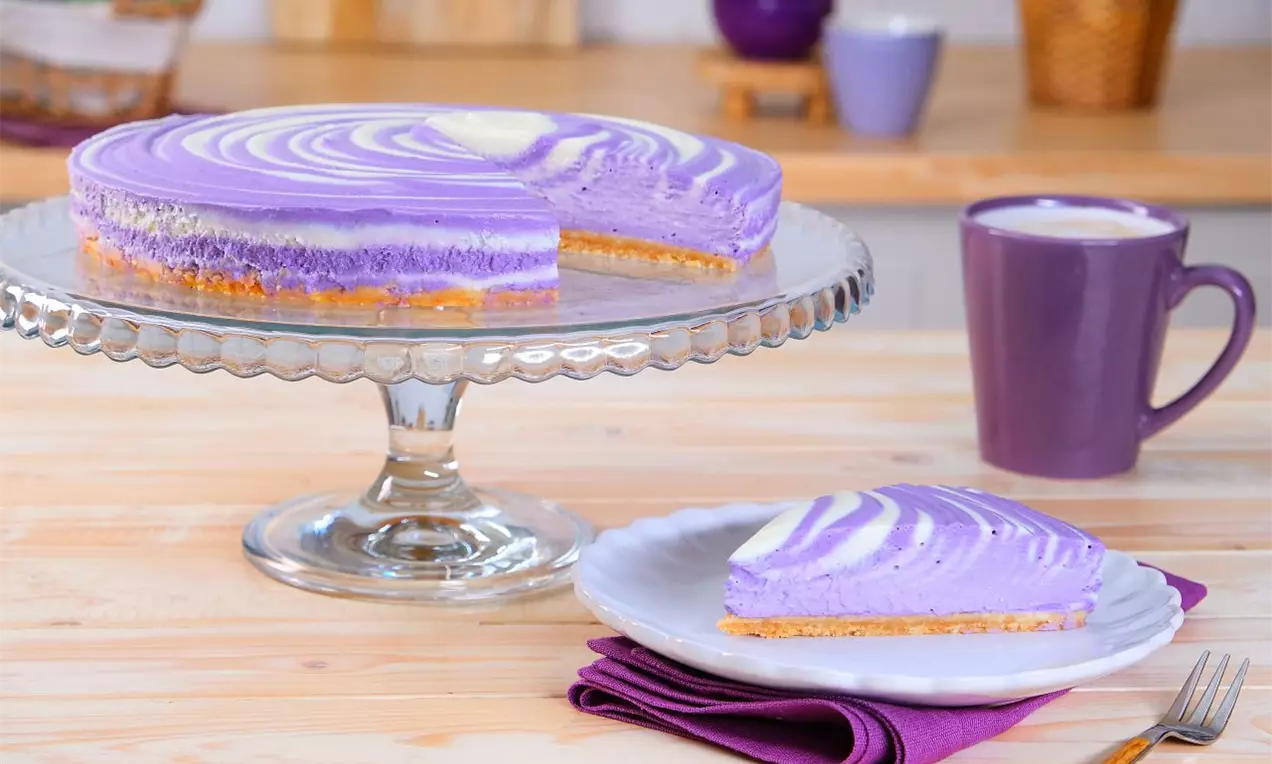 Ube cheesecake s hladkou fialovou náplňou s jemnými špirálami bielej farby, na zlatohnedom sušenkovom základe, servírovaný na sklenenom podnose