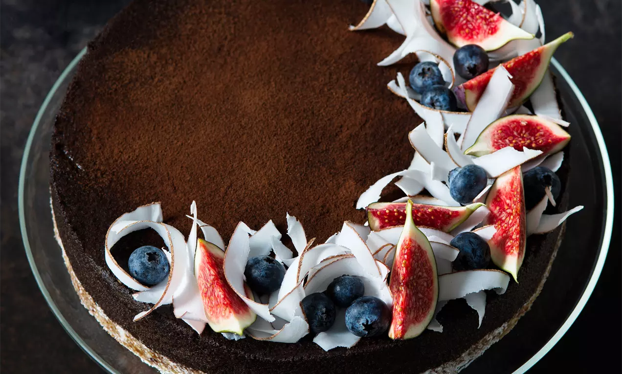 Nepečený okrúhly cheesecake s tmavohnedým karobovým povrchom, zdobený kokosovými hoblinami, čerstvými figami a modrými čučoriedkami na sklenenom podnose