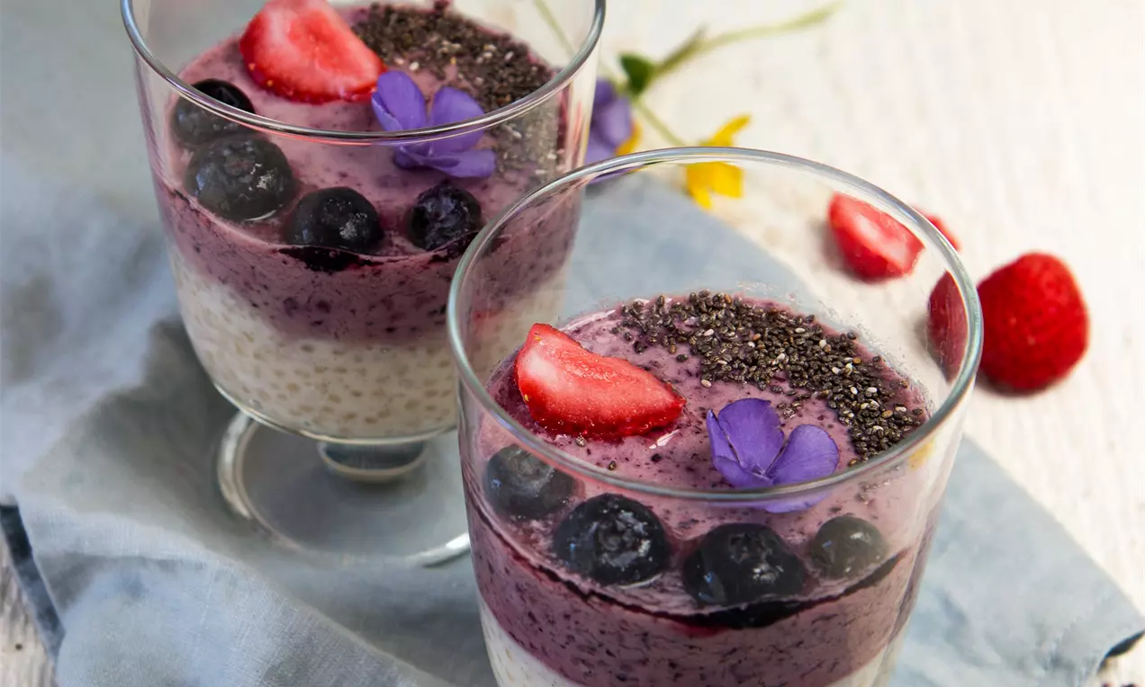 Vrstva čučoriedkového smoothie sýtej fialovej farby s celými čučoriedkami, jahodami, chia semienkami a fialovým kvetom v sklenenom pohári