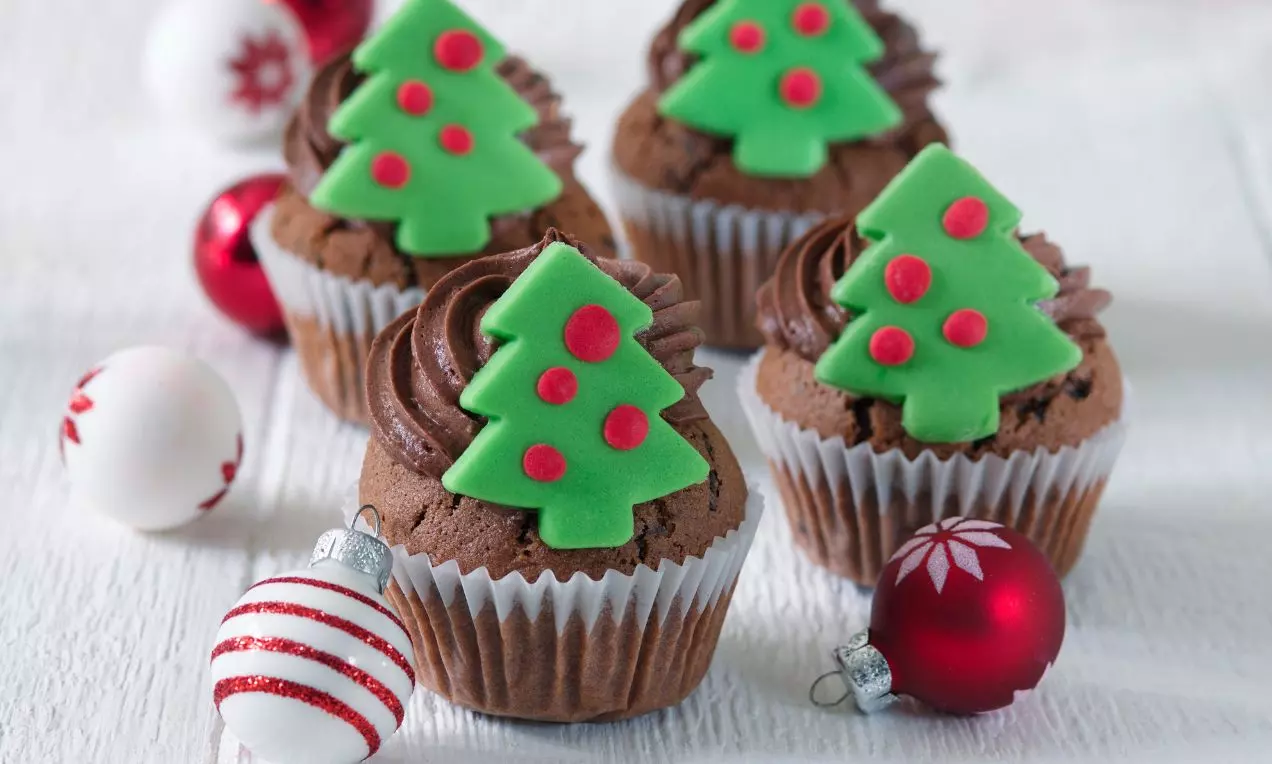 Čokoládové cupcakes s hladkým čokoládovým krémom, zelenými cukrovými stromčekmi a červenými guľkami, obklopené vianočnými ozdobami