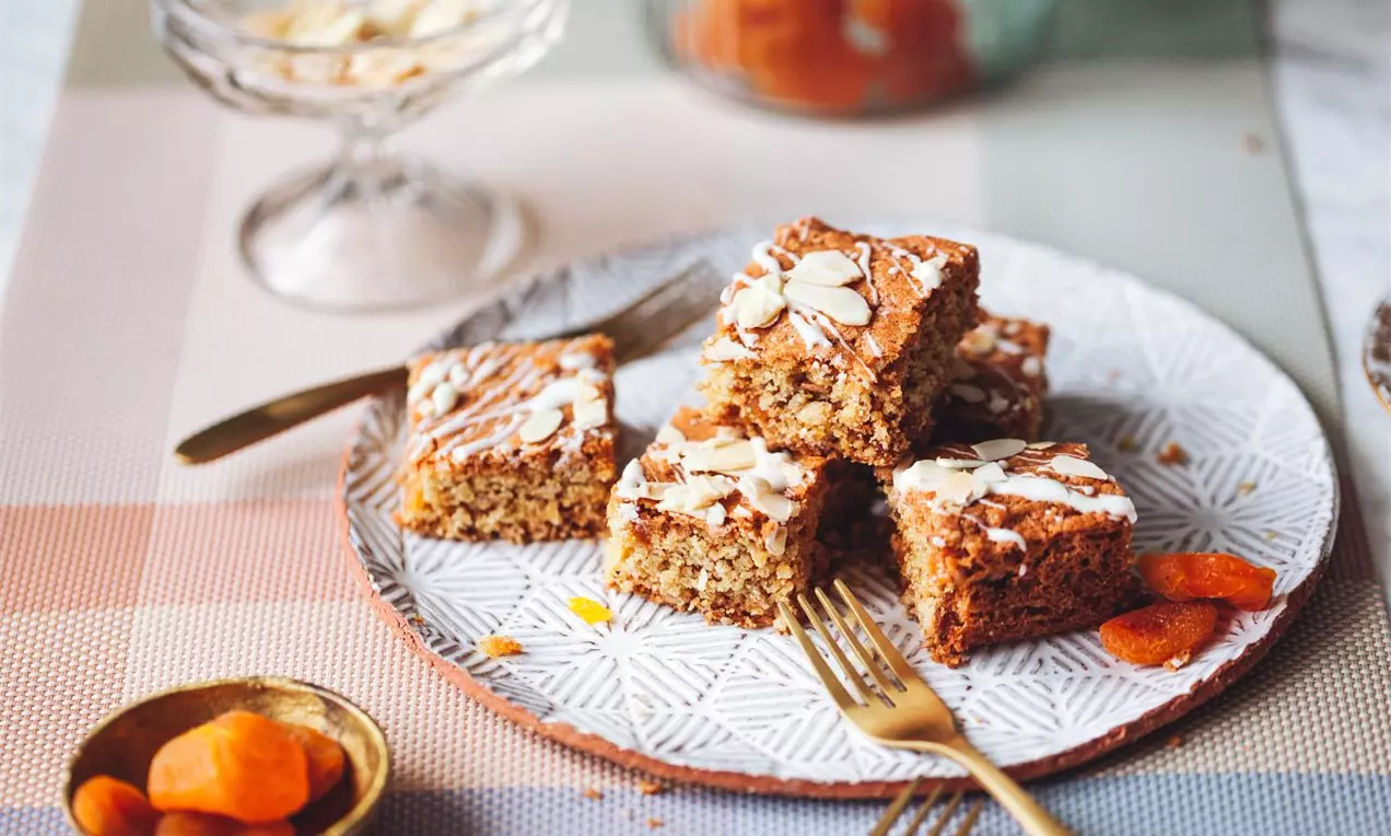 Blondie rezy servírované na sivom tanieri so zlatými príborami, ozdobené mandľovými lupienkami, bielou čokoládou a sušenými marhuľami