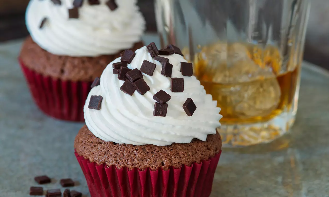 Čokoládové muffiny v červených košíčkoch, zdobené zvlneným kopčekom bielej šľahačky a čokoládovými kúskami na kovovom podnose pri pohári whisky
