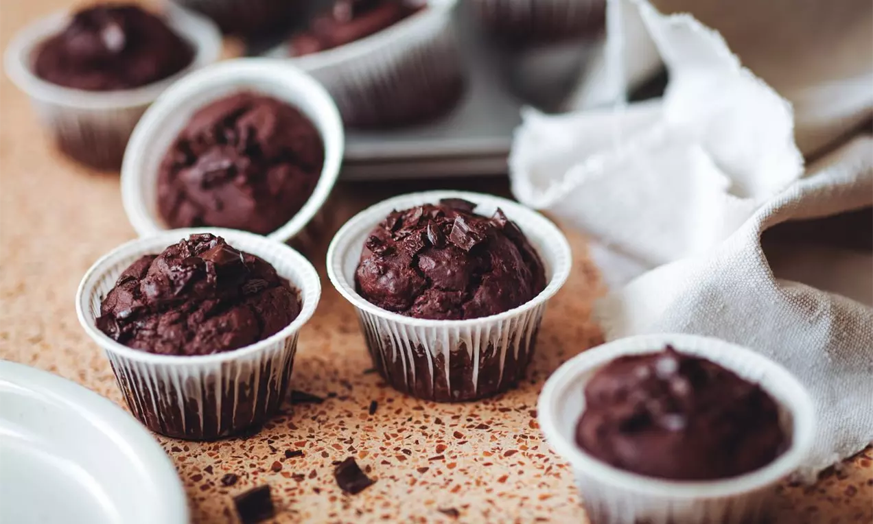 Čokoládovo hnedé muffiny s kúskami čokolády v bielych papierových košíčkoch na svetlom zrnite vzorovanom povrchu vedľa bielej látky
