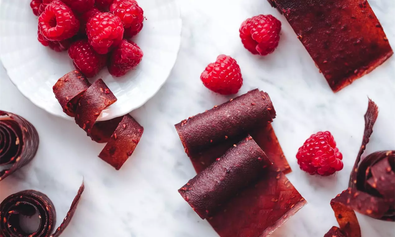 červené plátky z lisovaného ovocia srolované do rolky na bielej mramorovej pracovnej ploche s čerstvými malinami navôkol