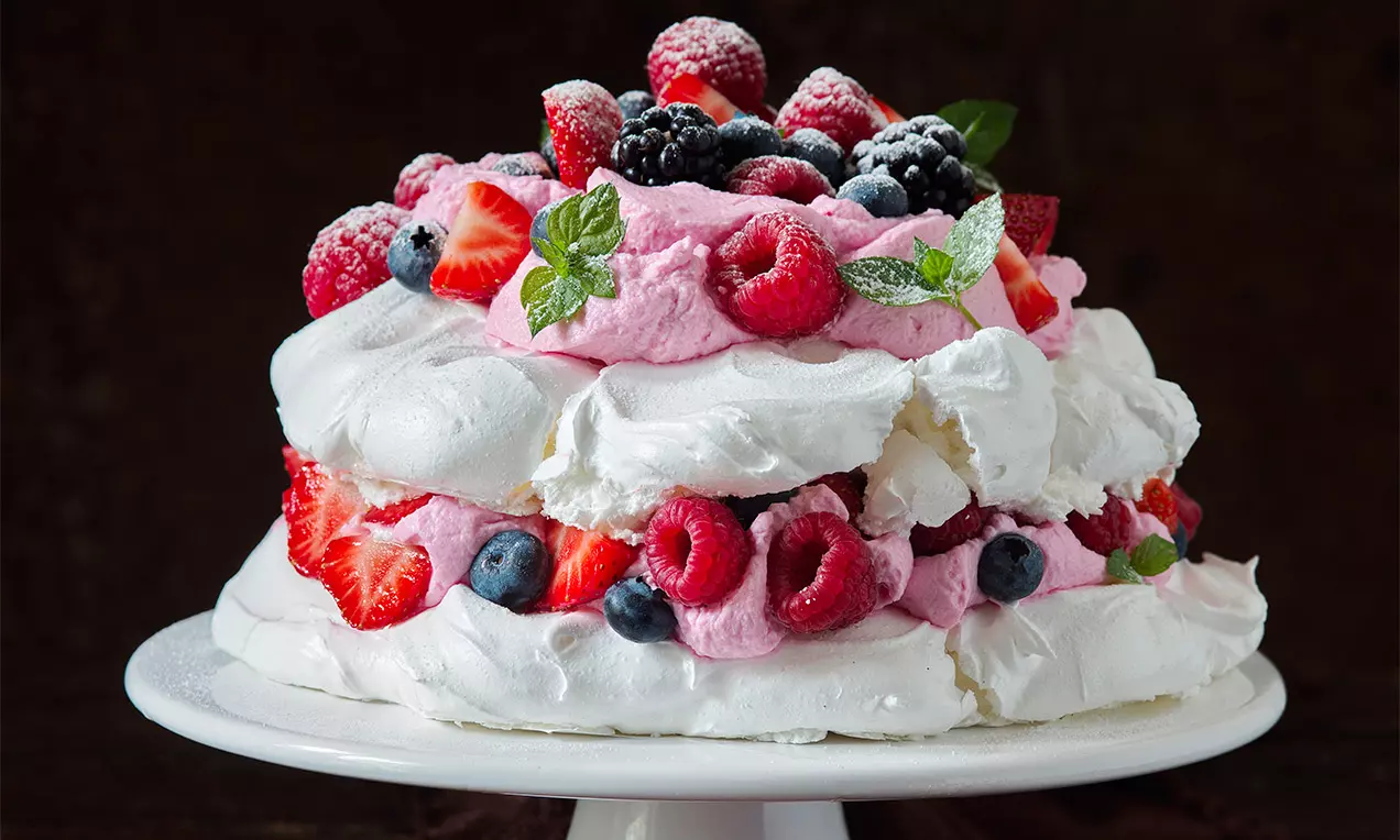 Dvojitá biela pavlova s nadýchanými vrstvami, ružovým krémom, čerstvými bobuľami a vetvičkou mäty na elegantnom bielom podnose