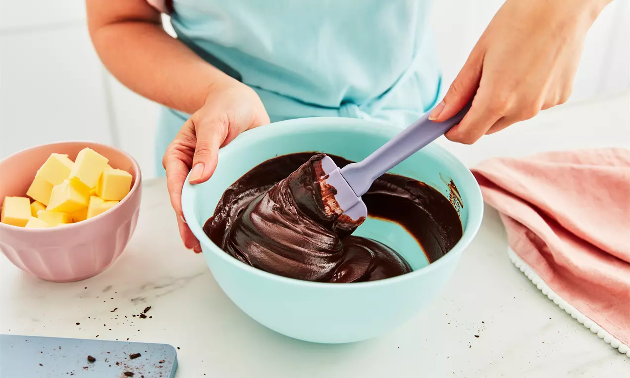 Ruky miešajú hladkú tmavú čokoládovú ganache pomocou silikónovej stierky v mise. Vedľa sú pripravené kocky masla, ktoré dodávajú ganache jemnú konzistenciu a lesklý vzhľad.