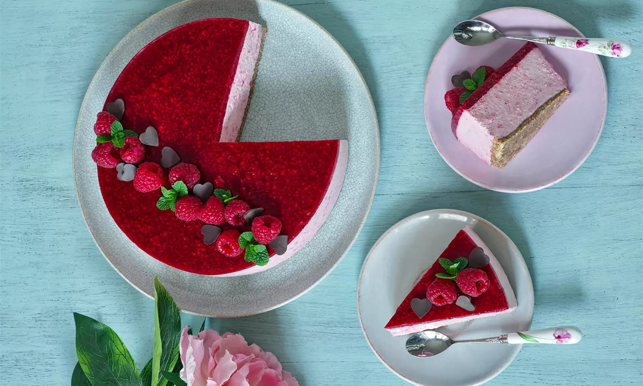 Malinový cheesecake s lesklou červenou polevou, ozdobený malinami, lístkami mäty a kúskami čokolády na bledomodrom a ružových tanieroch