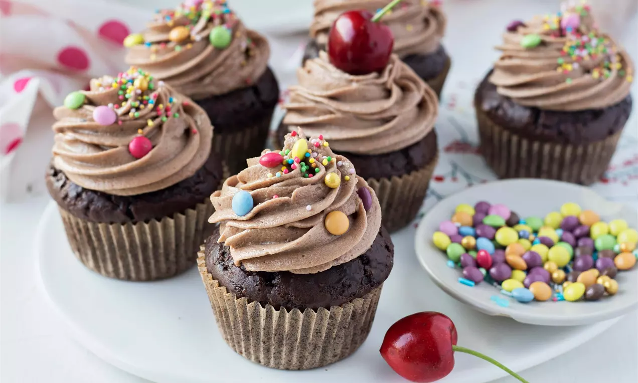 Čokoládové cupcakes s bohatou penovou polevou dozdobené farebným posypom a čerešňou na bielom tanieri vedľa misky s cukrovinkami