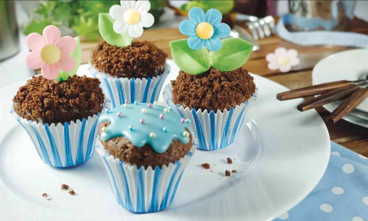 Čokoládové muffiny v modrobielych prúžkovaných košíčkoch s posypom z drveného cesta, zdobené farebnými kvetmi a lístkami na bielom tanieri  