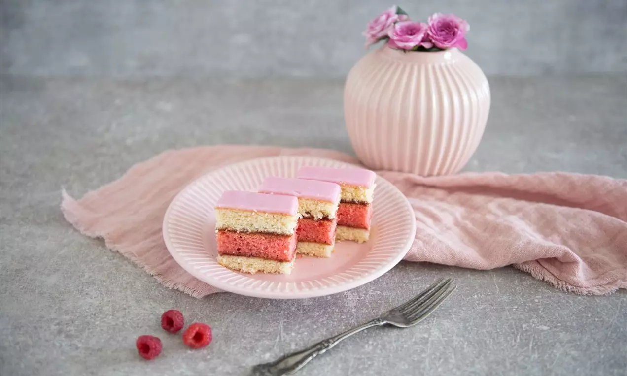 Malinový punčák tvorený vrstvami ružovej a svetlej piškótovej hmoty, preliaty ružovou polevou, na jemnom ružovom tanieri s malinami a vidličkou