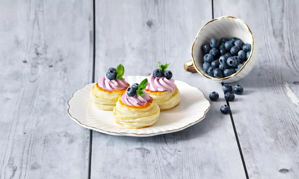 Nadýchané zlatisté pastry s jemným fialovým krémom, čučoriedkami a lístkami mäty na svetlom tanieri vedľa šálky plnej čerstvých čučoriedok