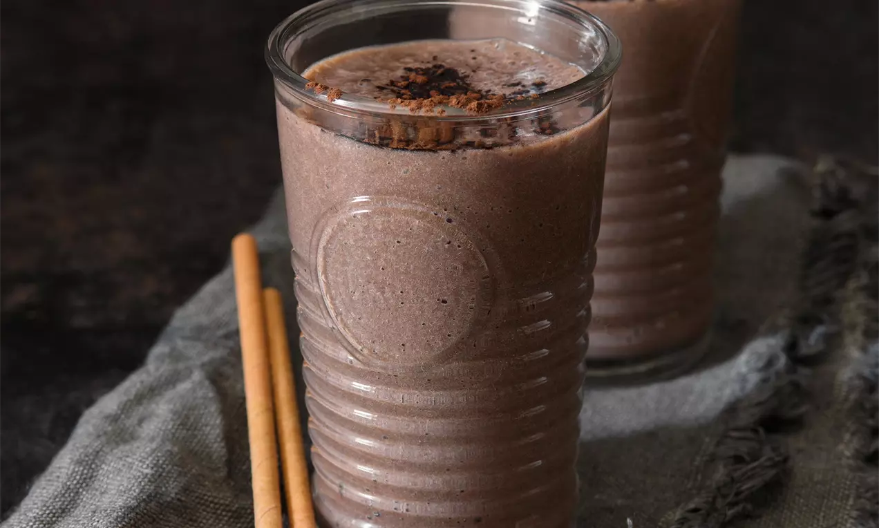 Hnedé banánovo-karobové smoothie v rebrovaných sklenených pohároch, hladké s jemnou penou na povrchu, posypané karobovým práškom na tmavom podklade