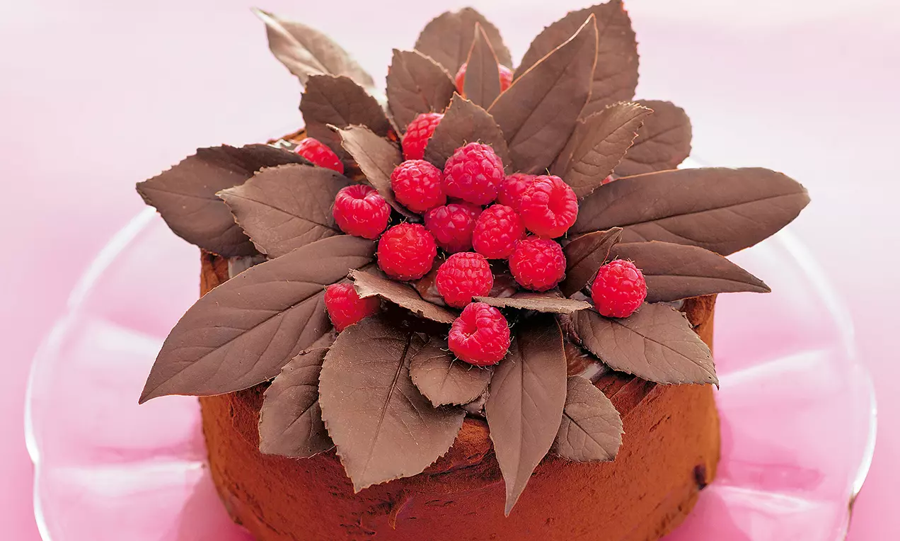 Torta zdobená tmavými čokoládovými listami a čerstvými malinami usporiadanými do kvetinového motívu na vrchnej časti čokoládovej torty.