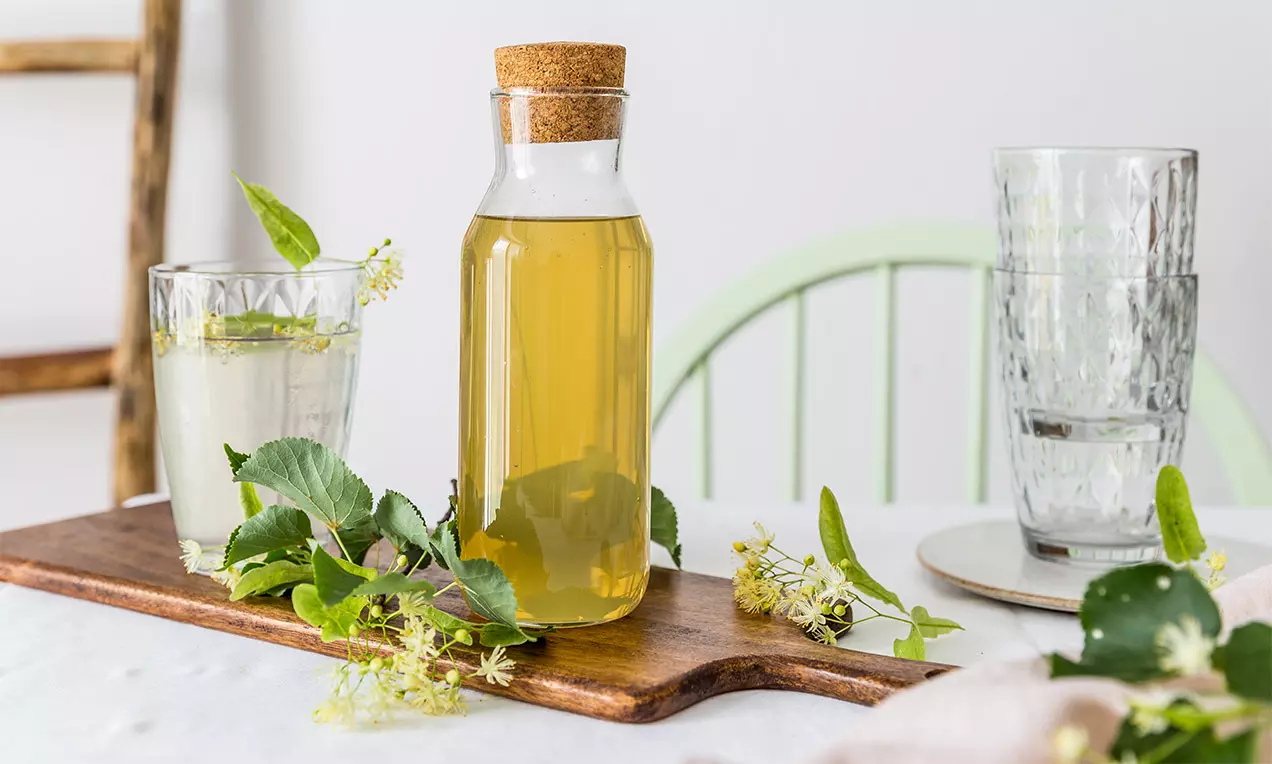 Svetložltý lipový sirup v priehľadnej fľaši s korkovým uzáverom, obklopený lipovými listami a kvetmi na drevenej doske
