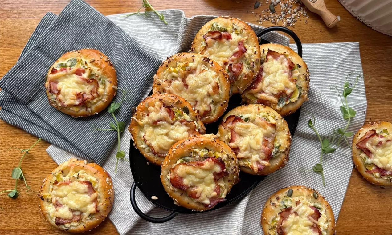 Kysnuté koláče so slaninou a syrom posypané semienkami, naaranžované na čiernom servírovacom podnose a ľanových utierkach na drevenom stole.