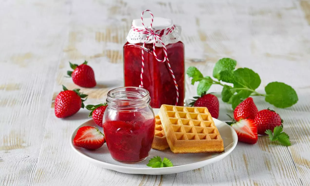 Pohár s jasne červeným jahodovým džemom vedľa čerstvých jahôd, lístkov mäty a zlatistých vaflí na svetlom drevenom podklade