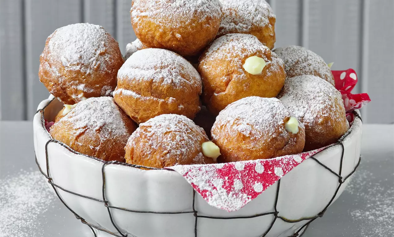 Zlatisté tvarohové šišky bohato posypané práškovým cukrom, naukladané na sebe v bielej drôtenej nádobe s červenou látkou