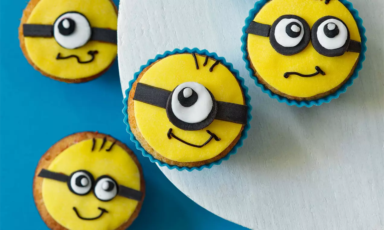 Banánové muffiny s jasne žltým vrchom zdobeným čiernymi a bielymi detailmi tvárí, uložené v modrých košíčkoch na modrobielom pozadí