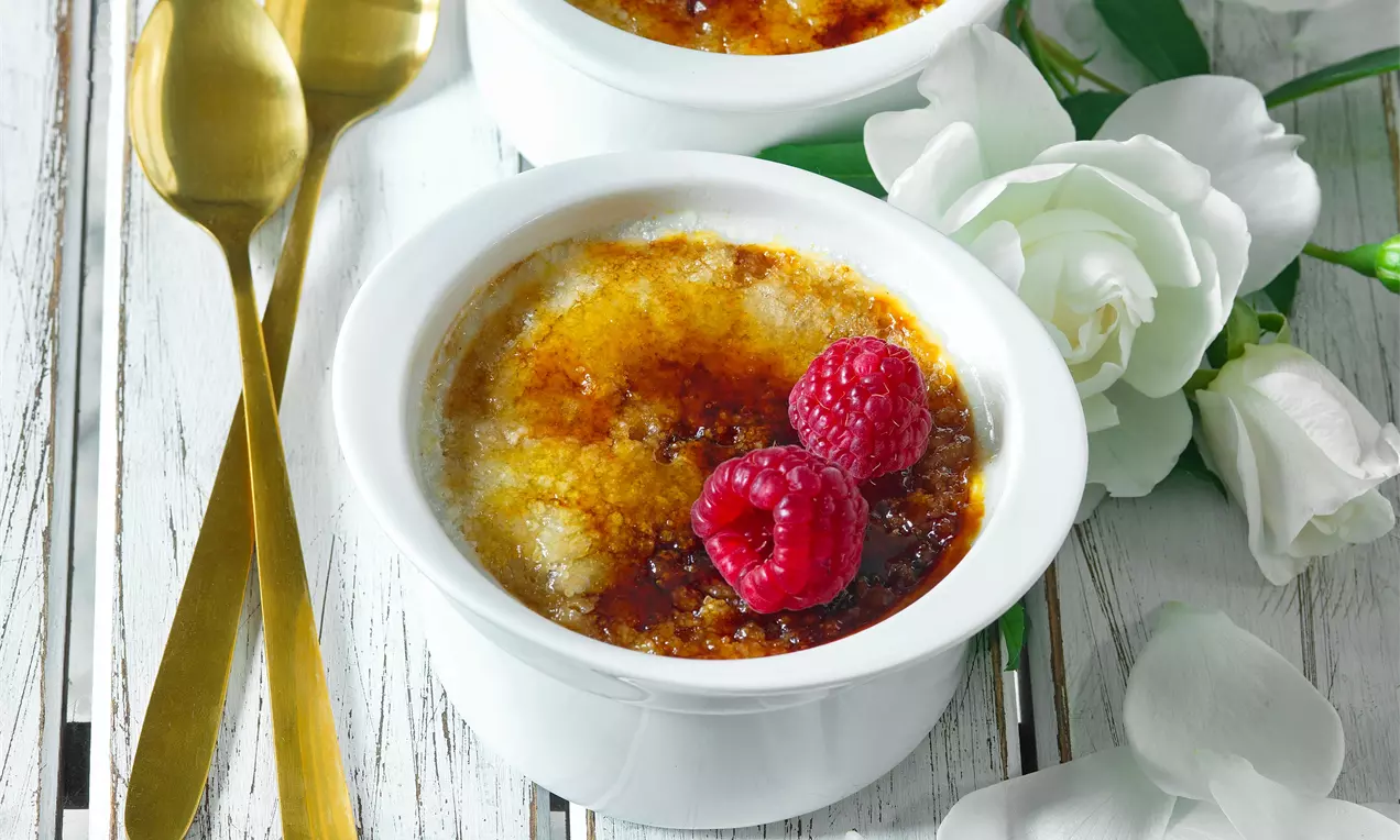 Crème brûlée s karamelovou krustou zlatej farby, jemný krém pod povrchom, ozdobený čerstvými malinami v bielej keramickej miske na dreve