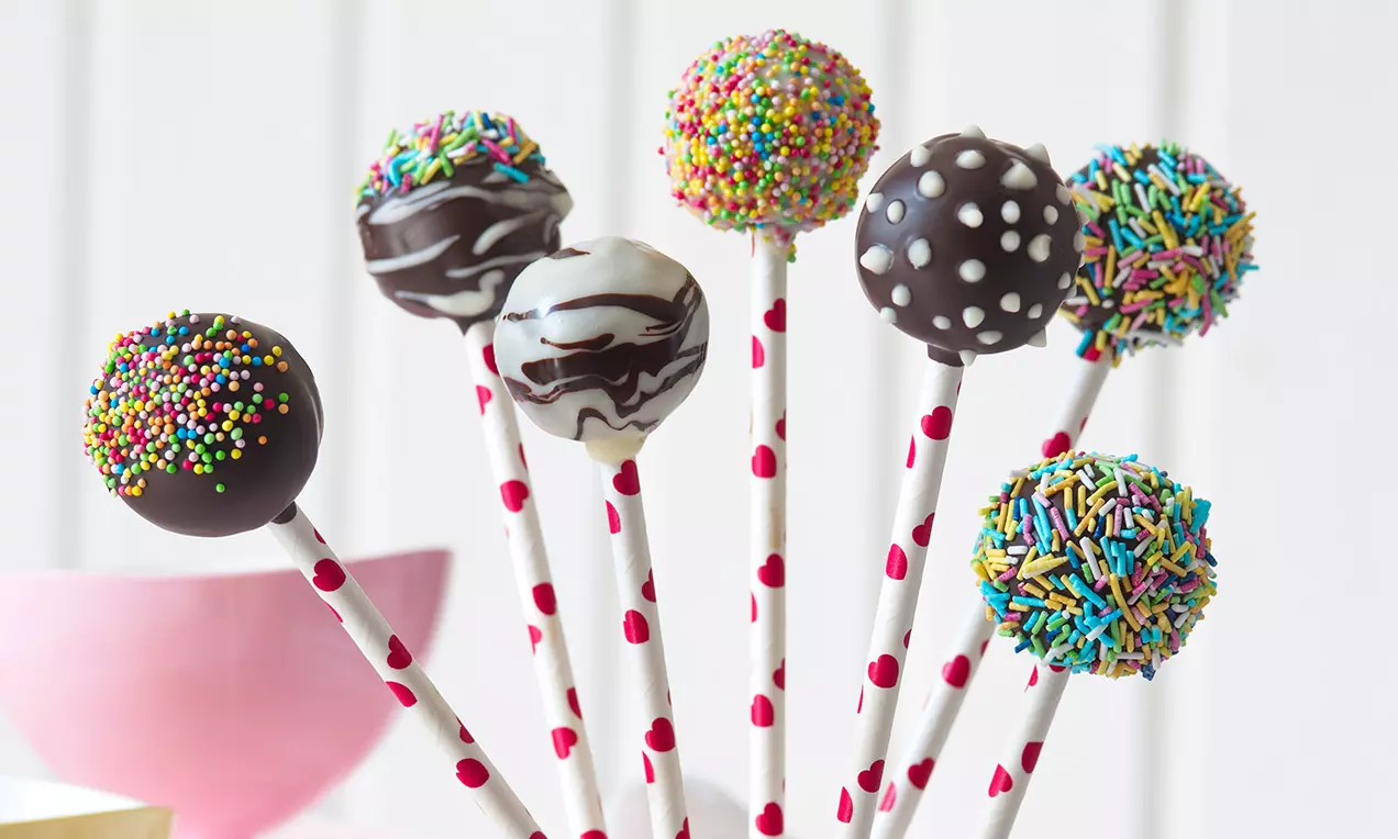 Perníkové cake pops na paličkách s bodkami a srdiečkami, ozdobené polevou, farebnými posýpkami a usporiadané vo veselom kytici v ružovej miske