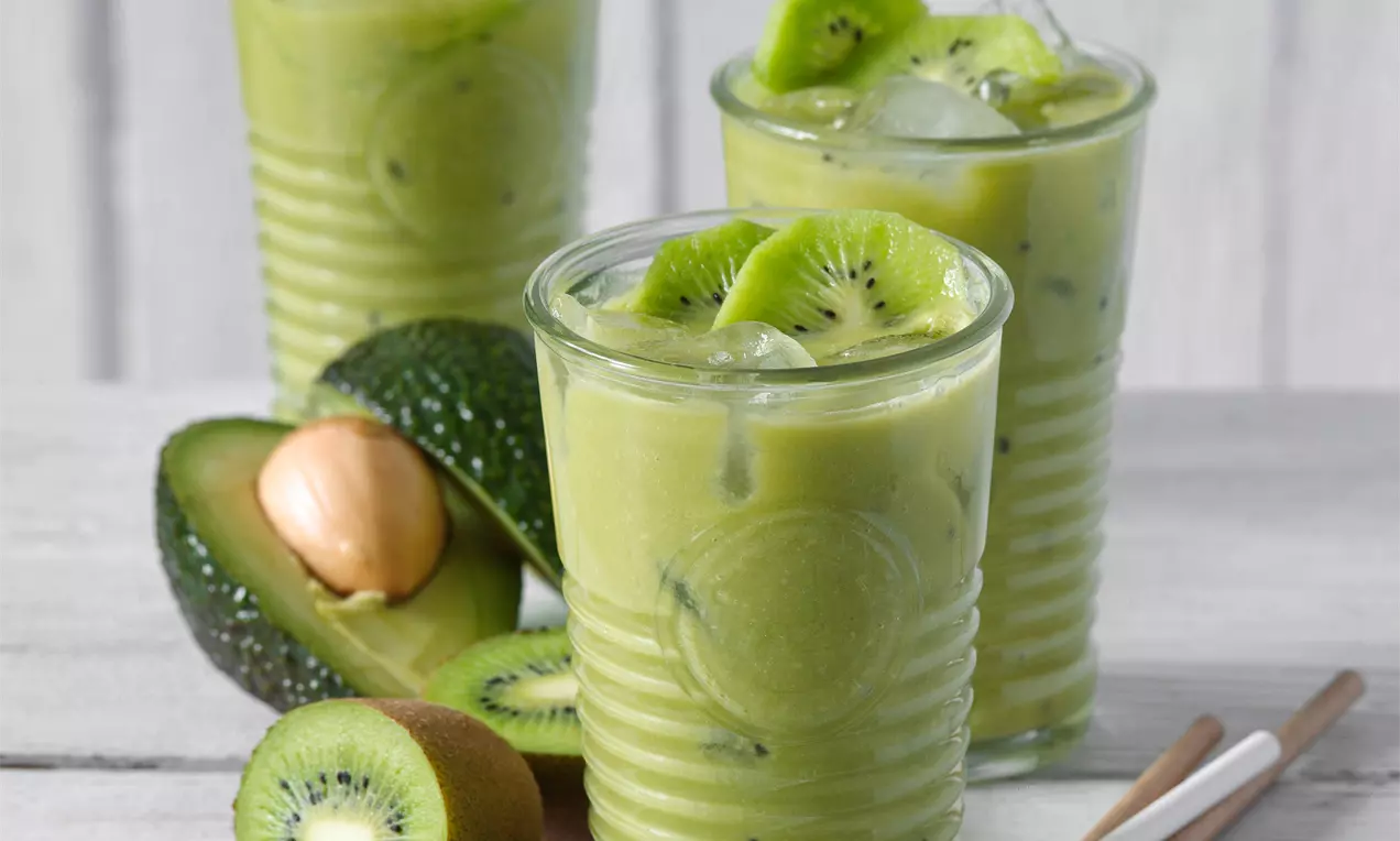 Svetlozelené smoothie v sklenených pohároch zdobené plátkami kivi, s kúskami avokáda vedľa na bledom drevenom povrchu