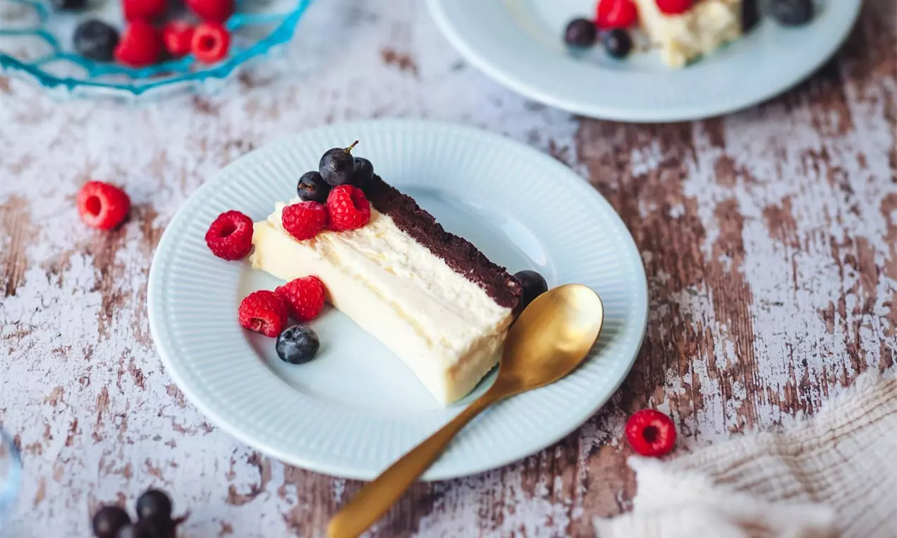 rez cheesecakeu s tmavým korpusom a bielym krémom na modrom dezertnom tanieriku, s okolím ozdobeným čerstvým lesným ovocím