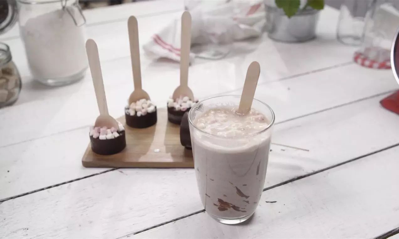 Kocky tmavej čokolády s posypom drobných bielych a ružových marshmallows na drevenej lyžičke vedľa pohára s krémovým nápojom na bielom stole