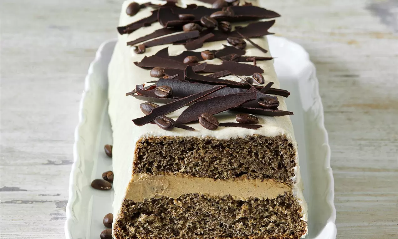 Biskupský chlebíček plnený kávovým krémom, poliaty bielkovou polevou a zdobený čokoládovými hoblinami a kávovými zrnkami, na bielom servírovacom tanieri a bielom drevenom stole.