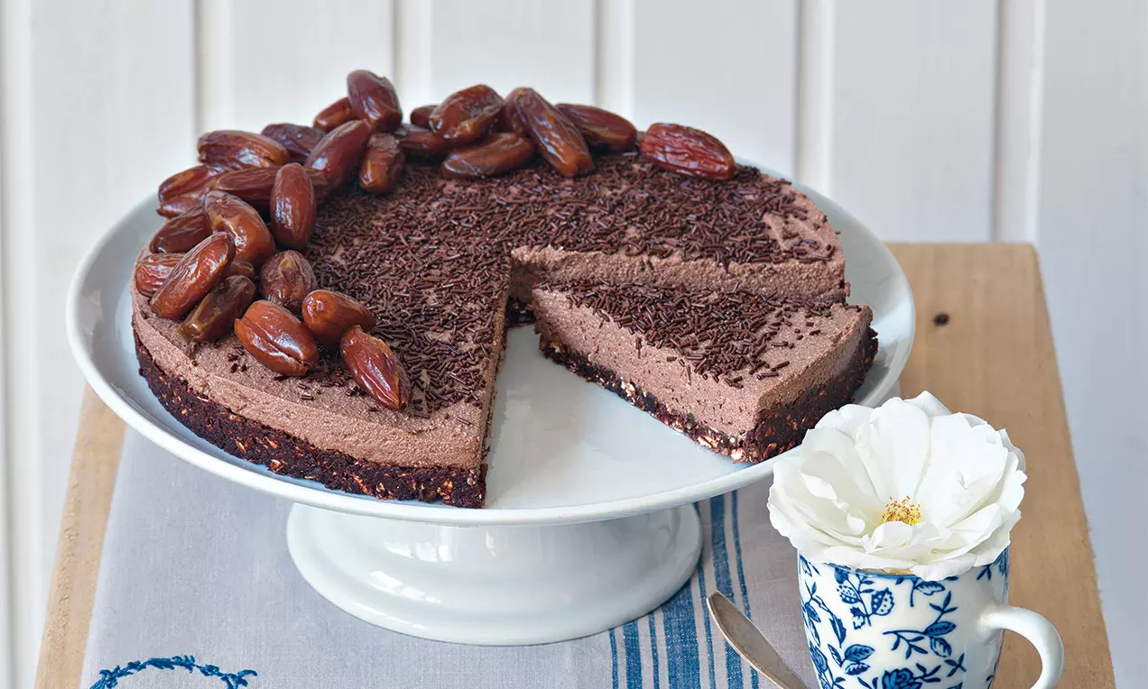 Cheesecake s kakaovým krémom na tmavom základe, hladký s jemnou štruktúrou, ozdobený datľami a čokoládovými hoblinkami na bielom stojane
