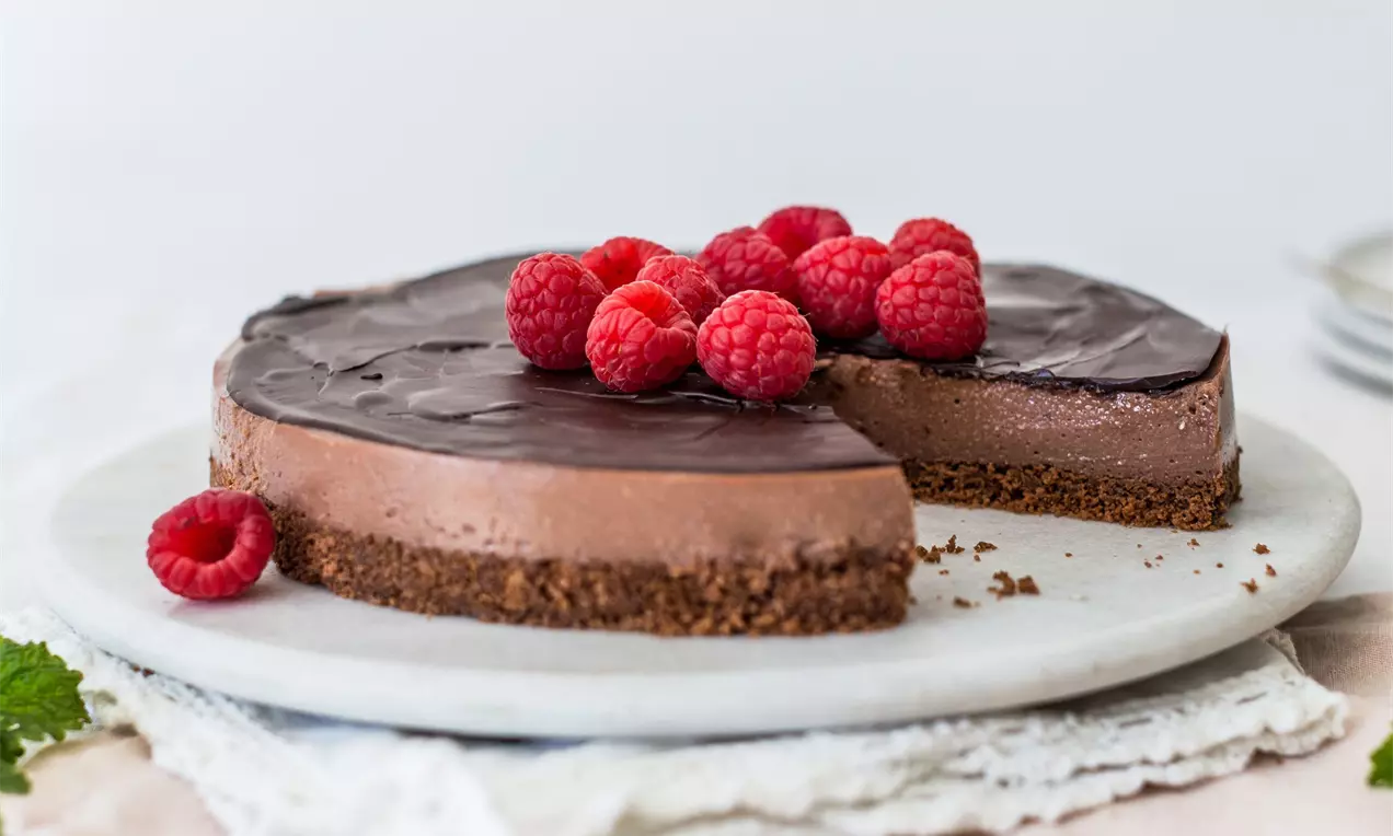 Čokoládový cheesecake s hladkým lesklým povrchom, kakaovým sušienkovým základom, ozdobený čerstvými malinami na bielom servírovacom podnose