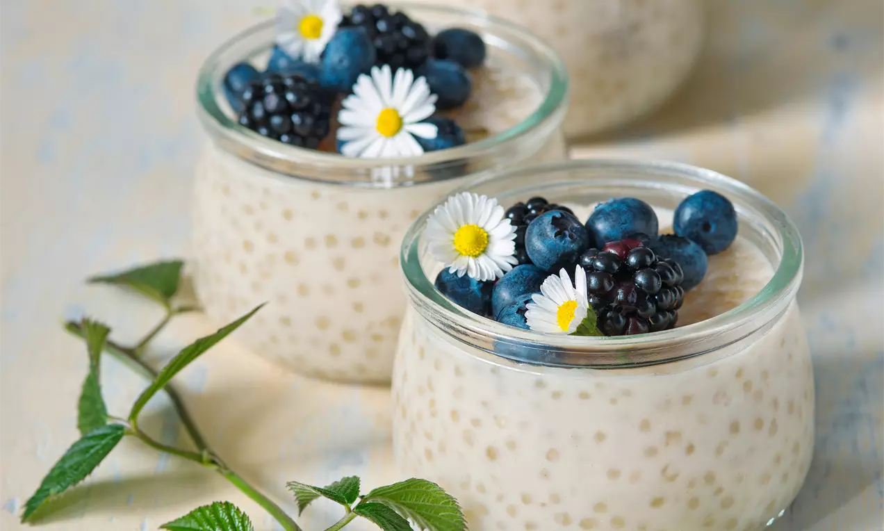 Krémový bielošedý tapiokový puding v sklenených pohároch, ozdobený čerstvými černicami, čučoriedkami a bielymi margarétkami na drevenom podklade