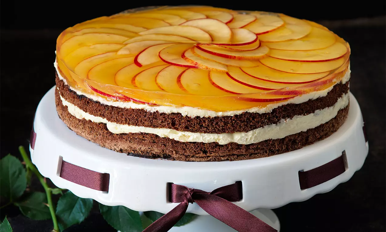 Jablková torta s vrstvou jemného krému medzi čokoládovými korpusmi, posiata tenkými plátkami jablka usporiadanými do kruhov na bielom podnose