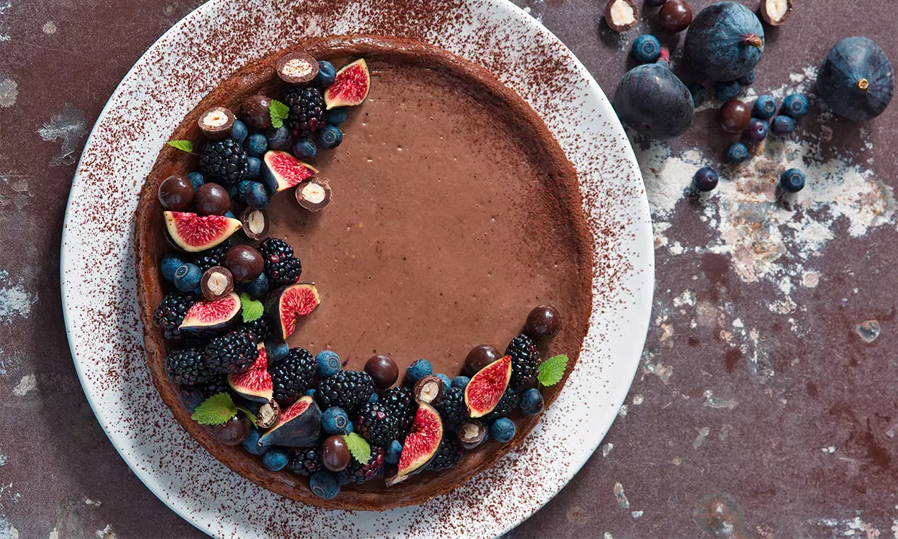 Kakaový cheesecake s hladkým hnedým povrchom, obložený čerstvými figami, čučoriedkami, černicami, lístkami mäty a kakaovým práškom na bielom tanieri