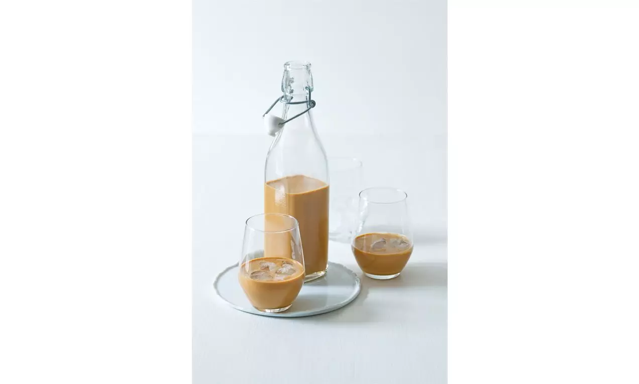 Sklenená fľaša s mandľovým likérom svetlohnedej farby vedľa dvoch pohárov s ľadovými kockami na bielom podnose položenom na svetlom stole