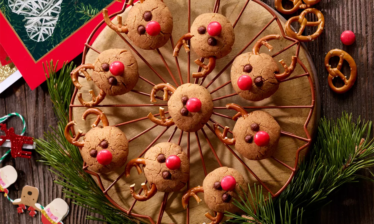 Krehké hnedé cookies v tvare sobích hláv s červenými nosmi, čokoládovými očami a preclíkovými parohami usporiadané na mriežke obklopenej vianočnými dekoráciami